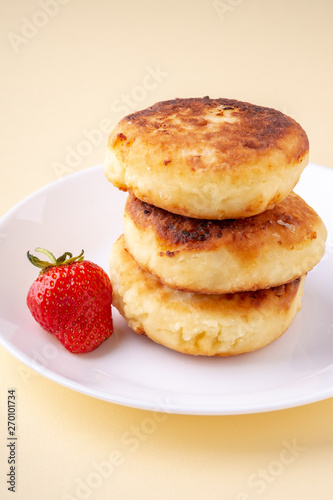 Cottage cheese pancakes with one strawberry on white plate on yellow background isolated angle view