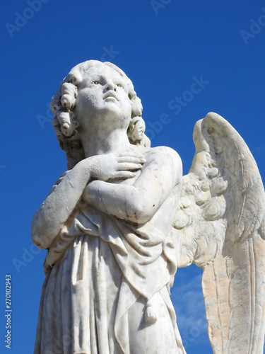 Scene in a graveyard  close-up of an old stone statue of an angel with arms crossed in the chest and looking at the sky. In the background  blue sky on a sunny day. Bottom view.