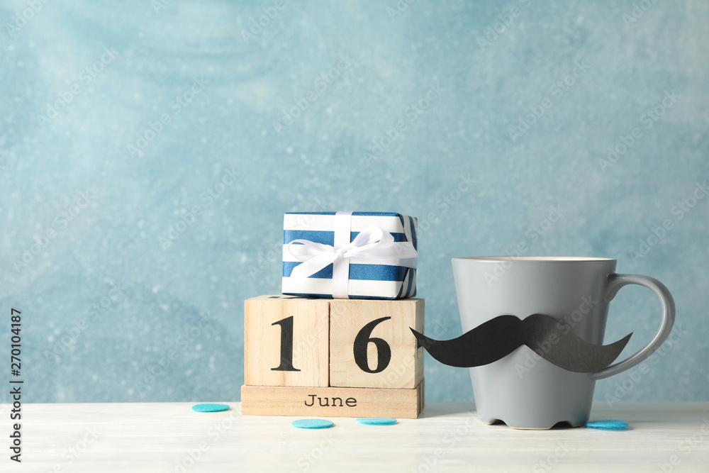 Obraz premium Cup with decorative mustache, gift box and wooden calendar on white table against blue background, space for text