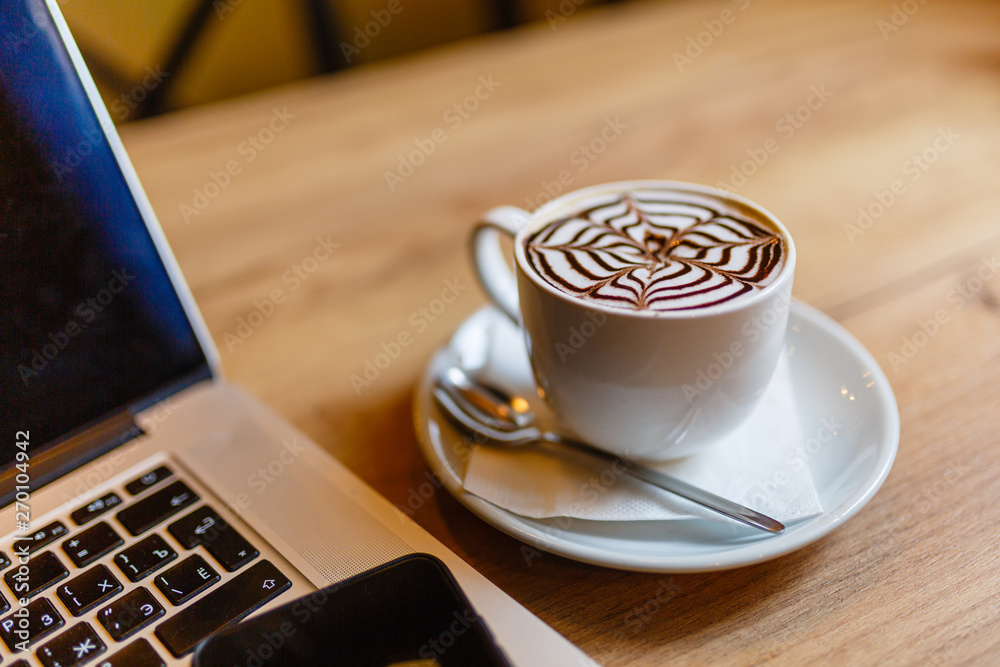 White Cup of cappuccino with pattern on the foam. Hot freshly brewed coffee in a white mug next to the smartphone and a laptop on a wooden table in the interior of the cafe. White Cup of cappuccino with pattern on the foam. Hot freshly brewed coffee in a white mug next to the smartphone and a laptop on a wooden table in the interior of the cafe.