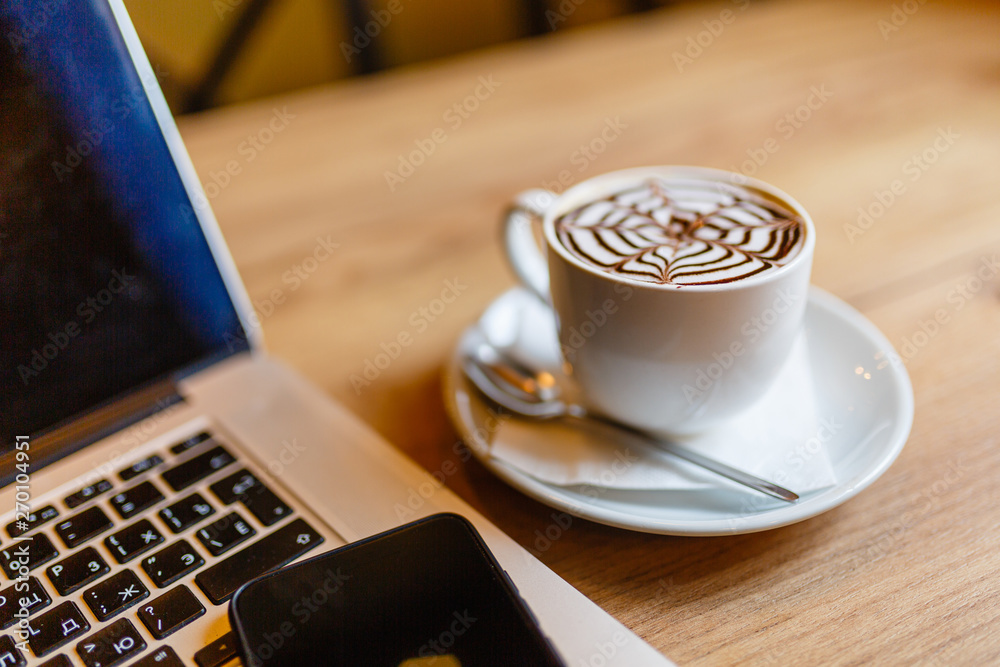 White Cup of cappuccino with pattern on the foam. Hot freshly brewed coffee in a white mug next to the smartphone and a laptop on a wooden table in the interior of the cafe. White Cup of cappuccino with pattern on the foam. Hot freshly brewed coffee in a white mug next to the smartphone and a laptop on a wooden table in the interior of the cafe.