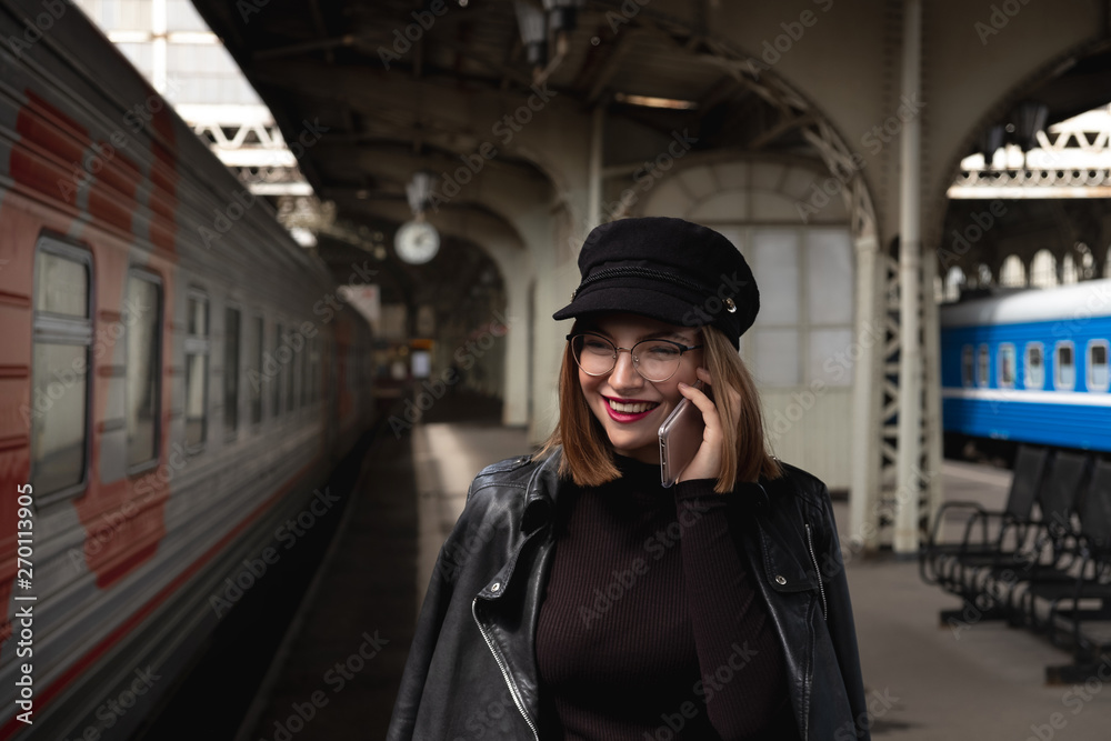 Attractive young woman millenial in black clothes and a hat at the railway station next to the train. Speaks on a mobile phone, smiles and waits for the train.