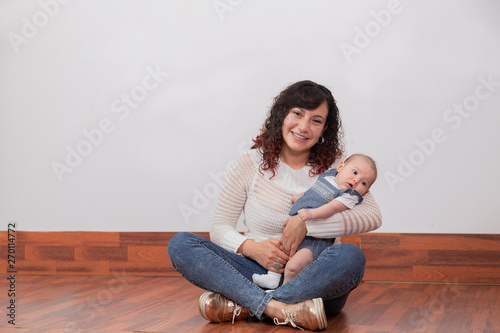 Mom sitting on the floor with her little daughter in her arms smiling and watching the camera both dressed in canvas - Hispanic young mom 