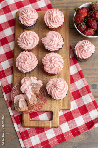 Homemade Strawberry Cupcakes on Wooden Cutting Board with one Open; Bowl of Fresh Strawberries Nearby; Red and White Checked Napkin on Wooden Table