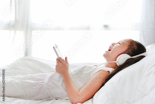 Happy little Asian girl using headphones listen music by smartphone while lying on bed in bedroom at home. © zilvergolf