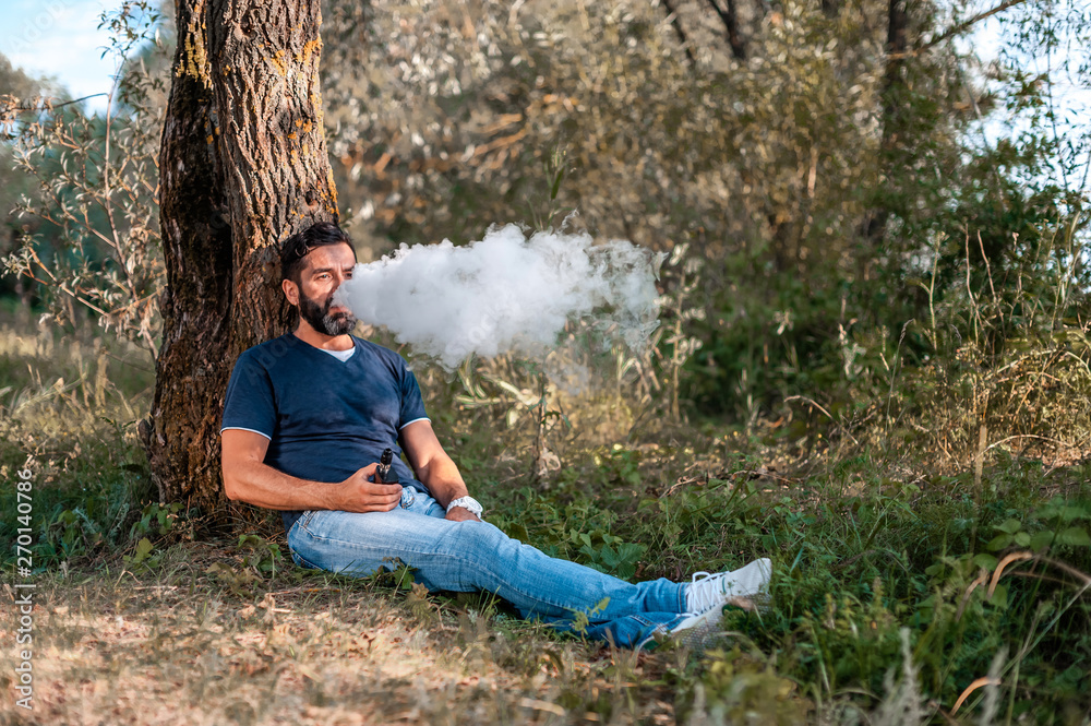 Bearded man blows up a couple an electronic cigarette on the forest ground. Getting rid of nicotine addiction.