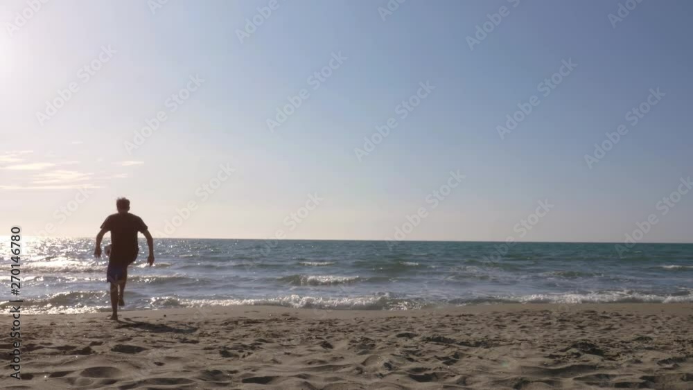 Young man in swimming trunks does tricks dribbling bouncing football on the sea shore at the beach at sunset silhouette Young man in swimming trunks does tricks dribbling bouncing football on the sea shore at the beach at sunset silhouette