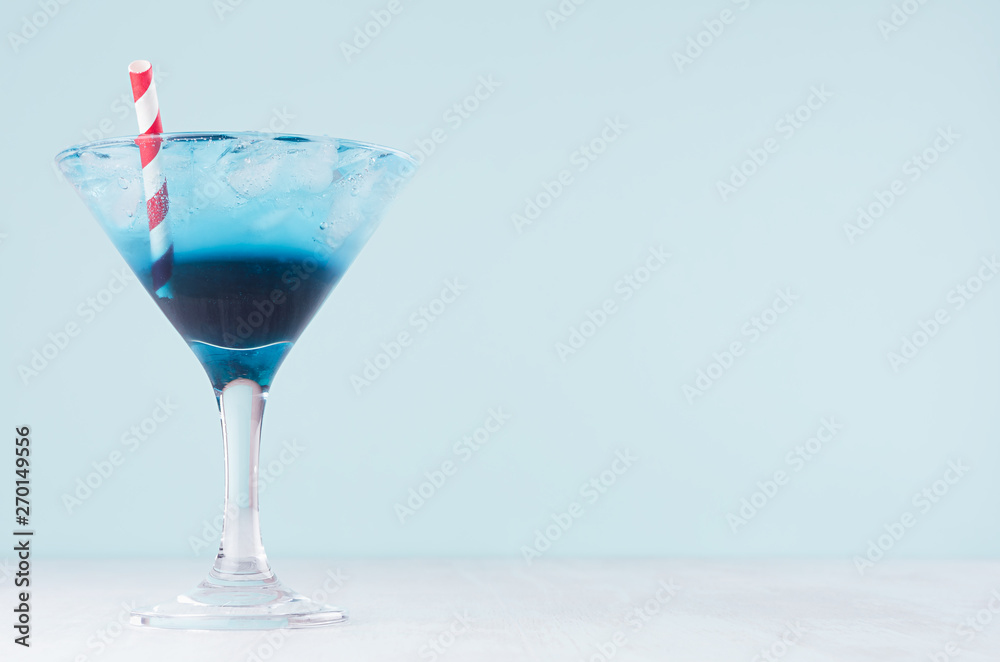 Cold tropical blue drink with ice, red straw in misted wineglass in modern pastel green interior on white wooden table.