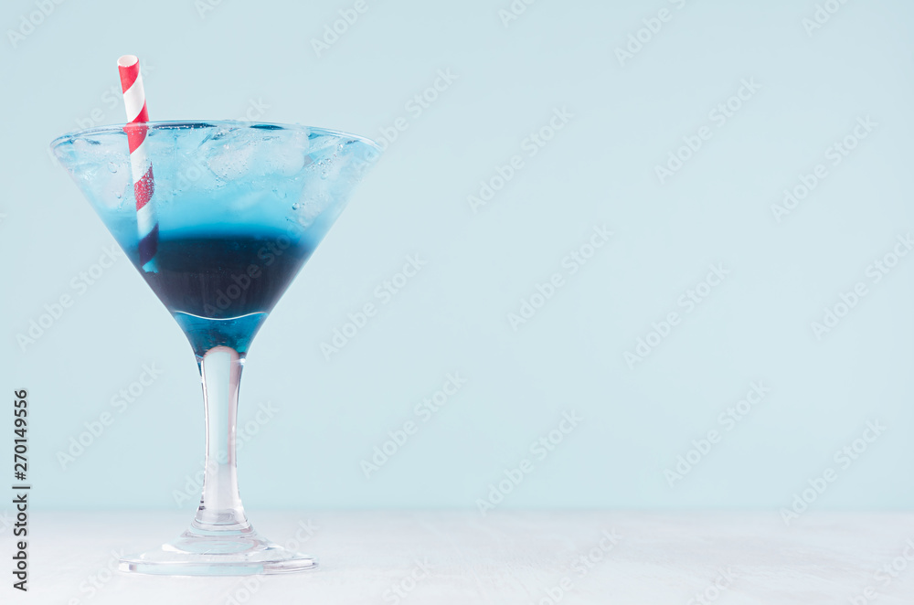 Cold tropical blue drink with ice, red straw in misted wineglass in modern pastel green interior on white wooden table. Cold tropical blue drink with ice, red straw in misted wineglass in modern pastel green interior on white wooden table.