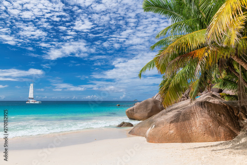 Ocean sunny beach with palms and a sailing boat in the turquoise sea on Seychelles island.