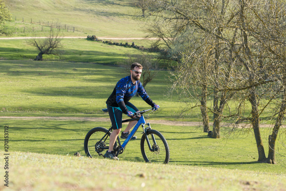 Cyclist in shorts and jersey on a modern carbon hardtail bike with an air suspension fork rides off-road on green hills near the forest Cyclist in shorts and jersey on a modern carbon hardtail bike with an air suspension fork rides off-road on green hills near the forest