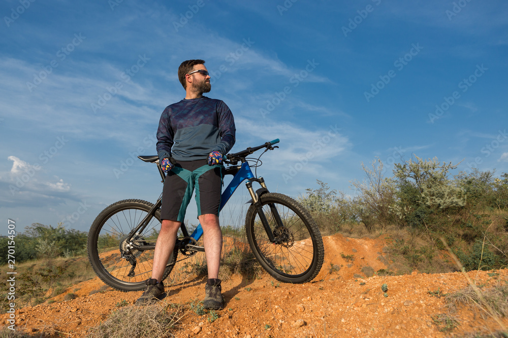 Obraz premium Cyclist in shorts and jersey on a modern carbon hardtail bike with an air suspension fork rides off-road on the orange-red hills at sunset evening in summer