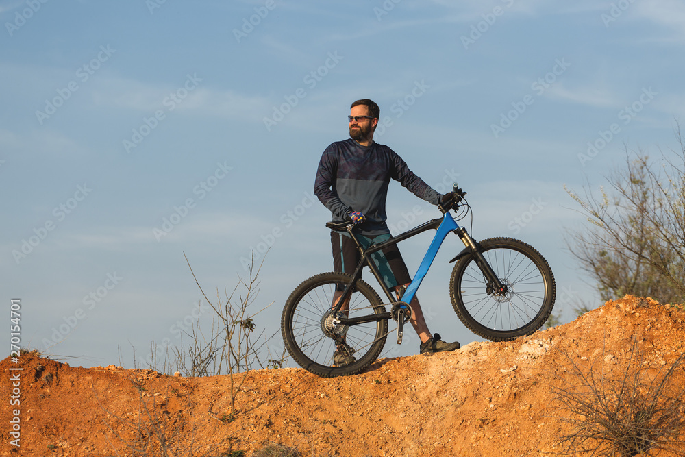 Cyclist in shorts and jersey on a modern carbon hardtail bike with an air suspension fork rides off-road on the orange-red hills at sunset evening in summer