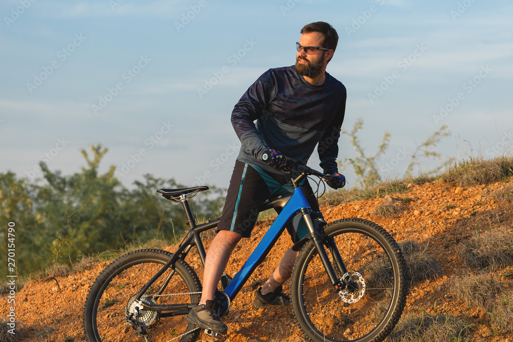 Fototapeta premium Cyclist in shorts and jersey on a modern carbon hardtail bike with an air suspension fork rides off-road on the orange-red hills at sunset evening in summer