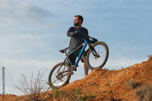 Cyclist in shorts and jersey on a modern carbon hardtail bike with an air suspension fork rides off-road on the orange-red hills at sunset evening in summer