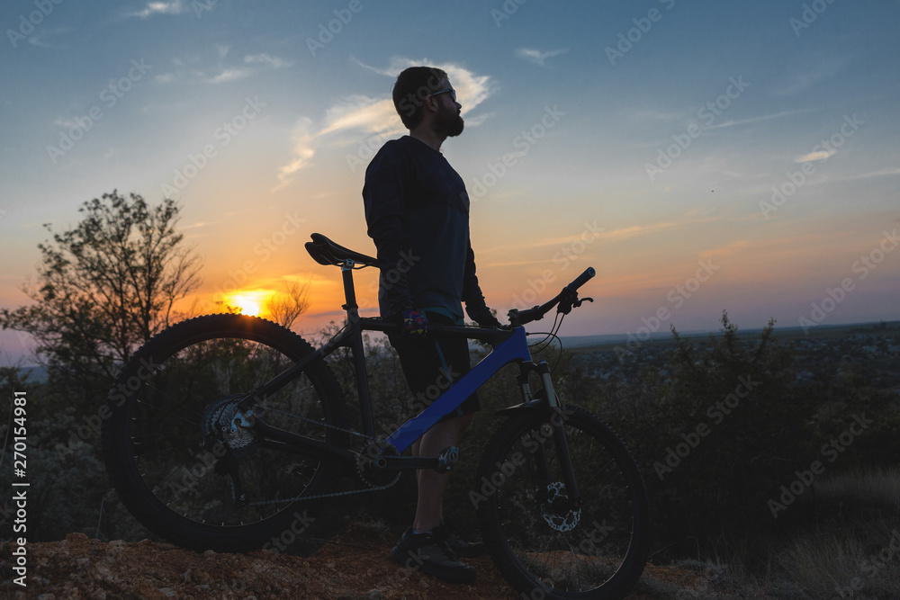 Cyclist in shorts and jersey on a modern carbon hardtail bike with an air suspension fork rides off-road on the orange-red hills at sunset evening in summer Cyclist in shorts and jersey on a modern carbon hardtail bike with an air suspension fork rides off-road on the orange-red hills at sunset evening in summer