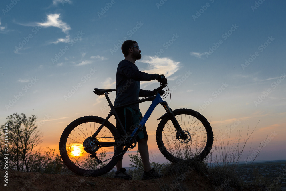 Cyclist in shorts and jersey on a modern carbon hardtail bike with an air suspension fork rides off-road on the orange-red hills at sunset evening in summer Cyclist in shorts and jersey on a modern carbon hardtail bike with an air suspension fork rides off-road on the orange-red hills at sunset evening in summer
