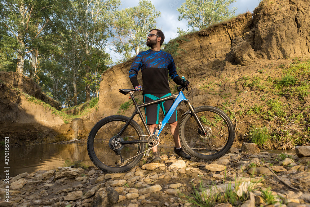 Obraz premium Cyclist in shorts and jersey on a modern carbon hardtail bike with an air suspension fork stands on the rocky shore of a mountain river