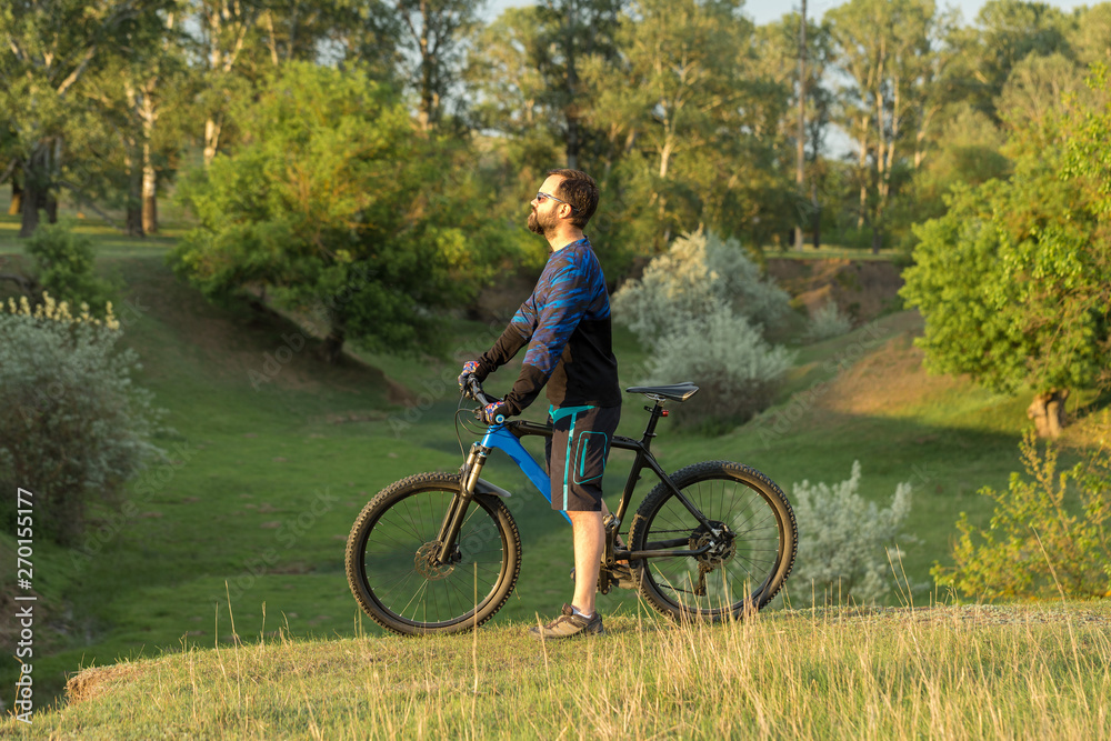 Cyclist in shorts and jersey on a modern carbon hardtail bike with an air suspension fork rides off-road on green hills near the forest Cyclist in shorts and jersey on a modern carbon hardtail bike with an air suspension fork rides off-road on green hills near the forest