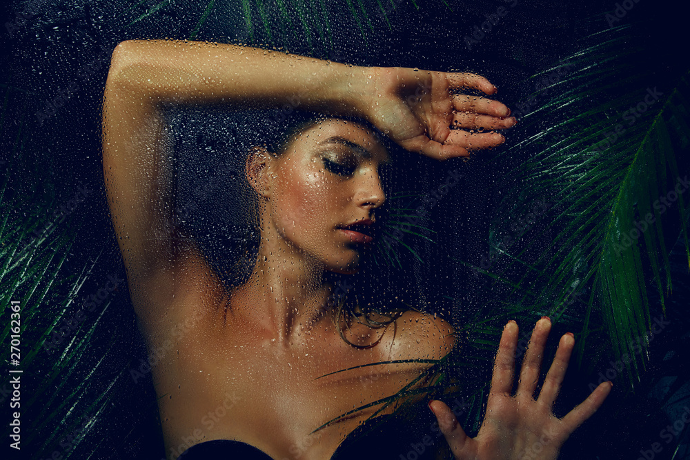 A beautiful, young, tanned girl with wet hair and wet skin is standing in the jungle in the rain. - obrazy, fototapety, plakaty 