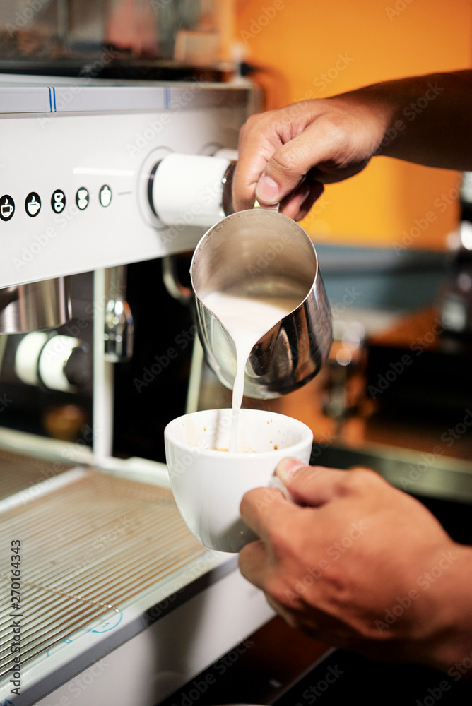 Close-up of male barista holding cup with coffee drink made with coffee machine and pouring milk from jug into this cup in cafe