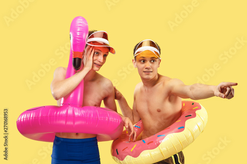 Two young men's half-length portrait isolated on yellow studio background. Smiling friends in caps with swimrings. Facial expression, summer, weekend, resort or vacation concept. Trendy colors.