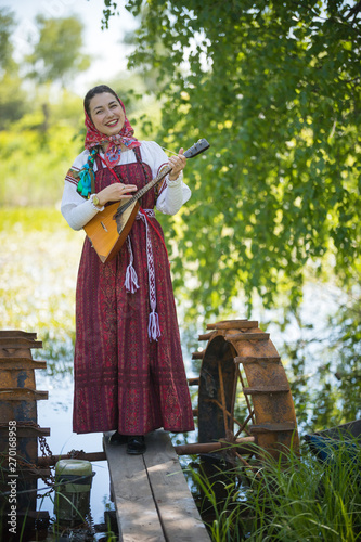 Young smiling woman in traditional russian clothes stands on a small bridge near the lake, and playing balalaika