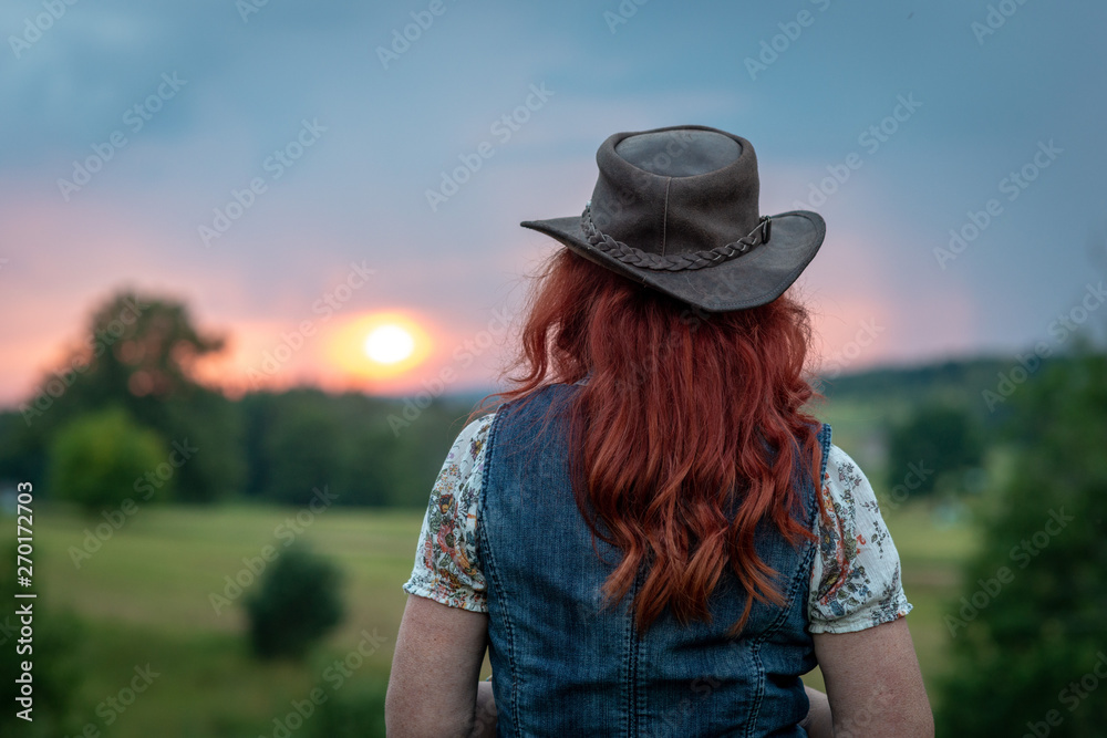 Woman with red hair and hat in head looks at sunset in the distance. Rear view.