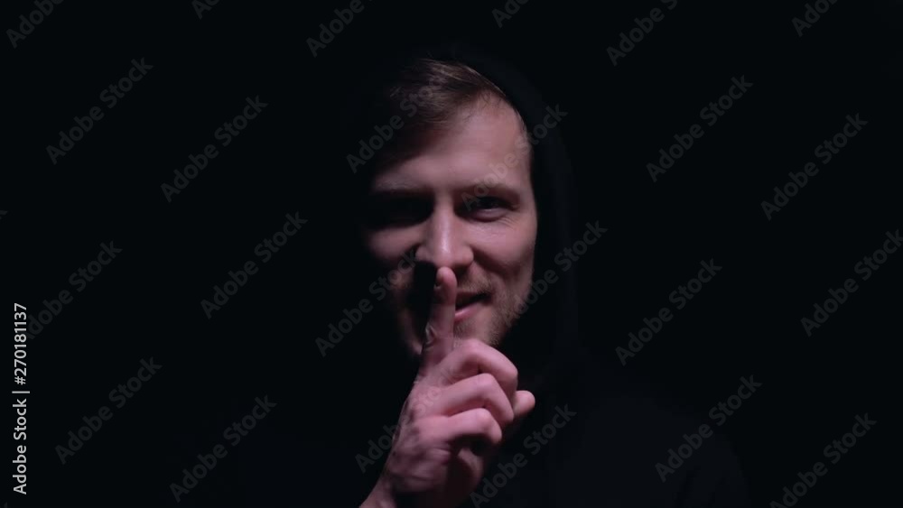 Cruel man in dark hood showing shh sign, intimidating victim, black background Cruel man in dark hood showing shh sign, intimidating victim, black background