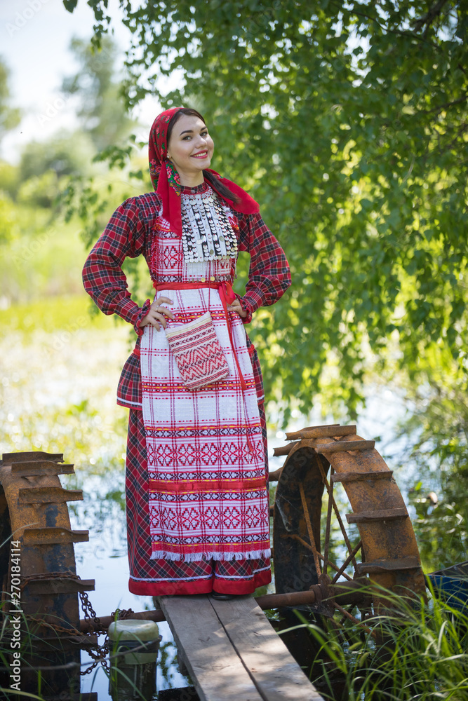 Young woman in scarf and traditional russian clothes stands on a small pier  near the lake and holds hands on a waist looking left Photos