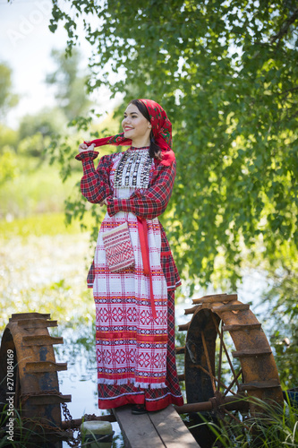 Attractive woman in scarf and traditional russian clothes stands on a small pier near the lake, looks right, posing for a photo