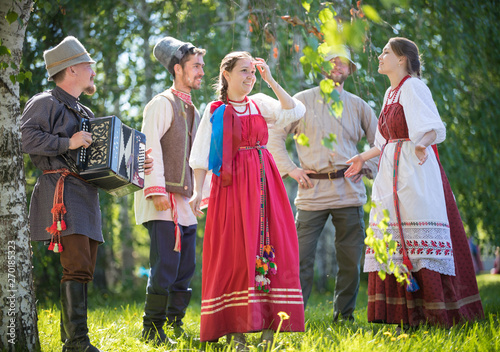 People in traditional Russian clothes communicate in the forest on the meadow - one of them plays music on accordion