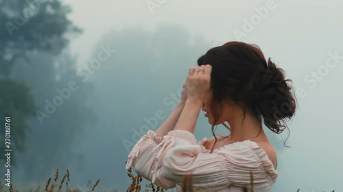 Beautiful girl in a gently pink dress, smiles, holds a blade of grass across the face, standing in a field on a summer day in nature and posing for the camera. Close-up.