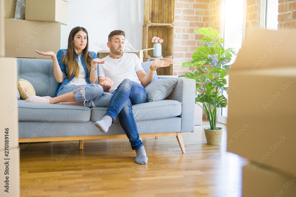 Young beautiful couple relaxing sitting on the sofa around boxes from moving to new house clueless and confused expression with arms and hands raised. Doubt concept. Young beautiful couple relaxing sitting on the sofa around boxes from moving to new house clueless and confused expression with arms and hands raised. Doubt concept.