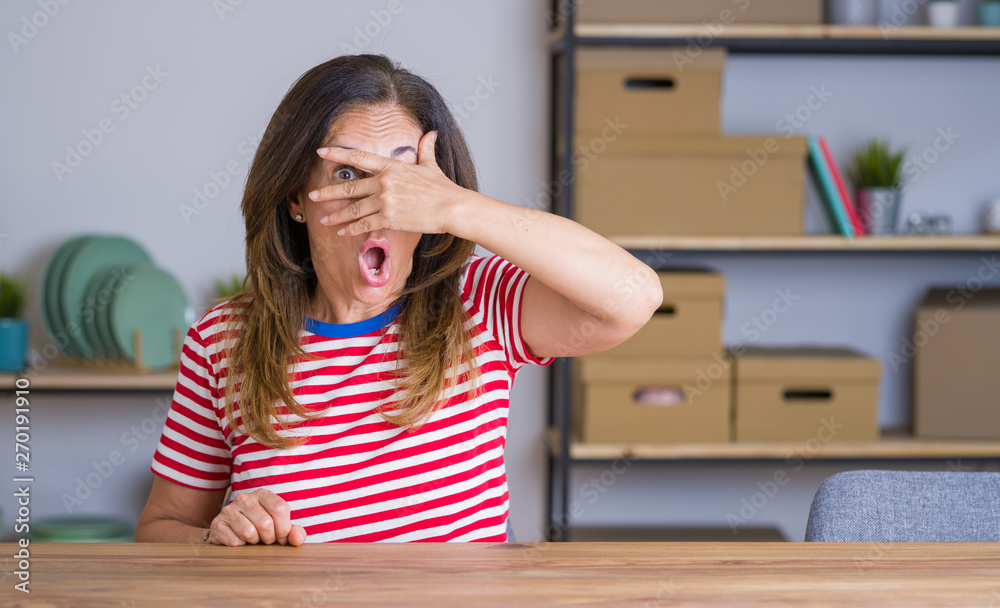 Middle age senior woman sitting at the table at home peeking in shock covering face and eyes with hand, looking through fingers with embarrassed expression. Middle age senior woman sitting at the table at home peeking in shock covering face and eyes with hand, looking through fingers with embarrassed expression.