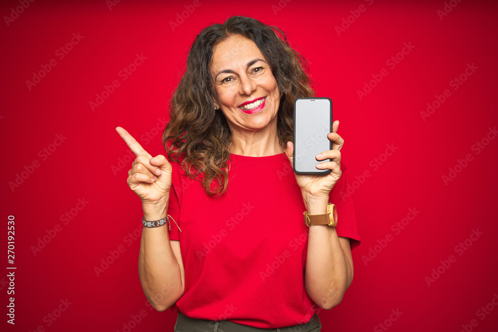 Middle age senior woman showing screen of smartphone over red isolated background very happy pointing with hand and finger to the side Middle age senior woman showing screen of smartphone over red isolated background very happy pointing with hand and finger to the side