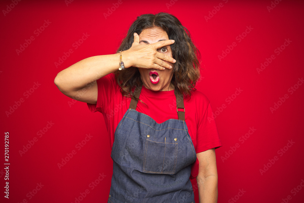 Middle age senior woman wearing apron uniform over red isolated background peeking in shock covering face and eyes with hand, looking through fingers with embarrassed expression. Middle age senior woman wearing apron uniform over red isolated background peeking in shock covering face and eyes with hand, looking through fingers with embarrassed expression.