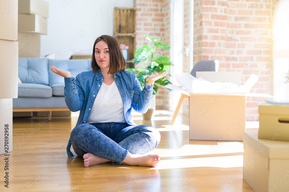 Young plus size woman sitting on the floor around cardboard boxes moving to a new home clueless and confused expression with arms and hands raised. Doubt concept. Young plus size woman sitting on the floor around cardboard boxes moving to a new home clueless and confused expression with arms and hands raised. Doubt concept.