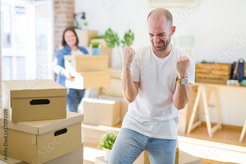Young couple arround cardboard boxes moving to a new house, bald man standing at home very happy and excited doing winner gesture with arms raised, smiling and screaming for success
