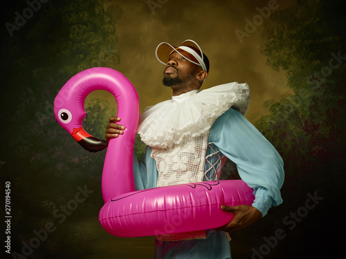 Young man as a medieval grandee or nobleman in a swimring as a pink flamingo on dark studio background. Portrait in retro costume. Human emotions, comparison of eras and facial expressions concept.