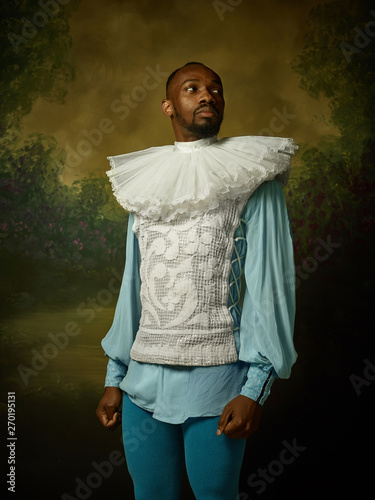 Young man as a medieval grandee or nobleman on dark studio background. Looking serious. Portrait of male model in retro costume. Human emotions, comparison of eras and facial expressions concept.