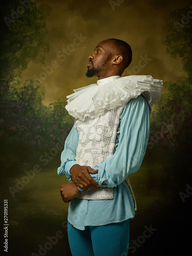 Young man as a medieval grandee or nobleman on dark studio background. Looking serious. Portrait of male model in retro costume. Human emotions, comparison of eras and facial expressions concept.
