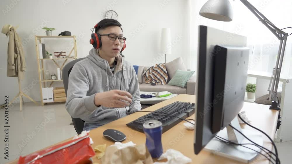 asian teenage boy nervous using computer playing game finger fast on keyboard. college student on summer holiday break lifestyle concept. handsome chinese man in earphones losing upset drink beverage