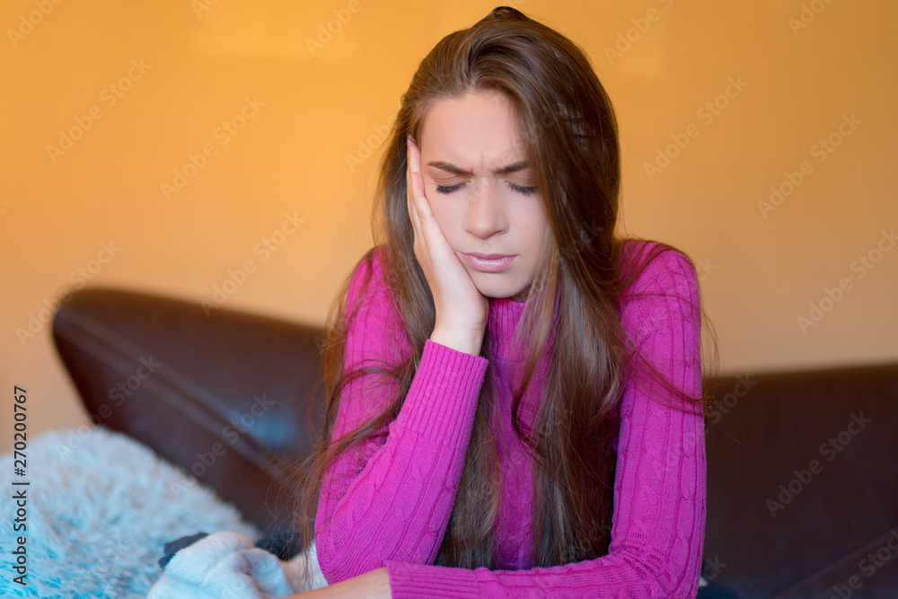 Toothache young woman touching her cheek and keeping eyes closed while sitting on the couch at home.