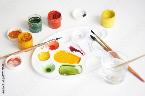 On a white background there is a still life with art supplies: gouache paints of different colors, brushes, palette and a glass of water to dilute the paint.
