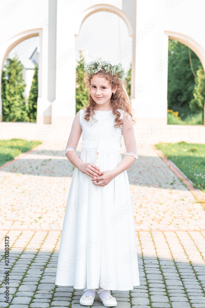 Portrait of cute little girl on white dress and wreath on first holy communion background church gate. first communion girl