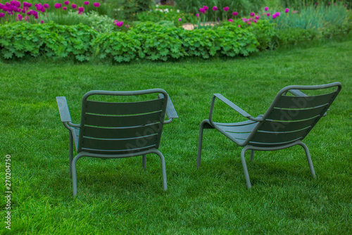 green chairs in the garden, dark green garden chairs for two on the green grass in front of flowers, spring relaxation in nature