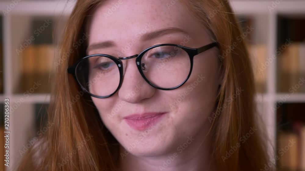 Closeup portrait of young redhead attractive caucasian female student in glasses looking at camera smiling with shyness in the college library Closeup portrait of young redhead attractive caucasian female student in glasses looking at camera smiling with shyness in the college library