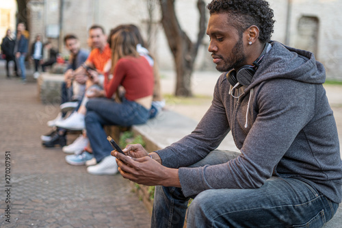 An African American guy playing alone with his smartphone. On the back a blurred group of friends having fun together. Racism and racial discrimination concept.