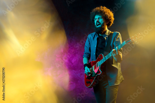 Hipster man with curly hair with red guitar in neon lights. Rock musician is playing electrical guitar.